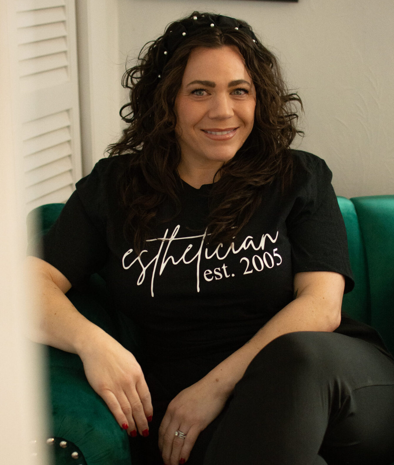 Jessica sitting in a velvet green chair in her suite, wearing a black tee and legs crossed.